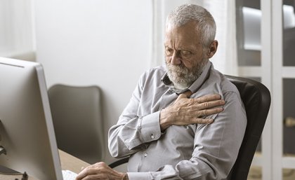 Man sitting at a laptop clutching at this chest with one hand 